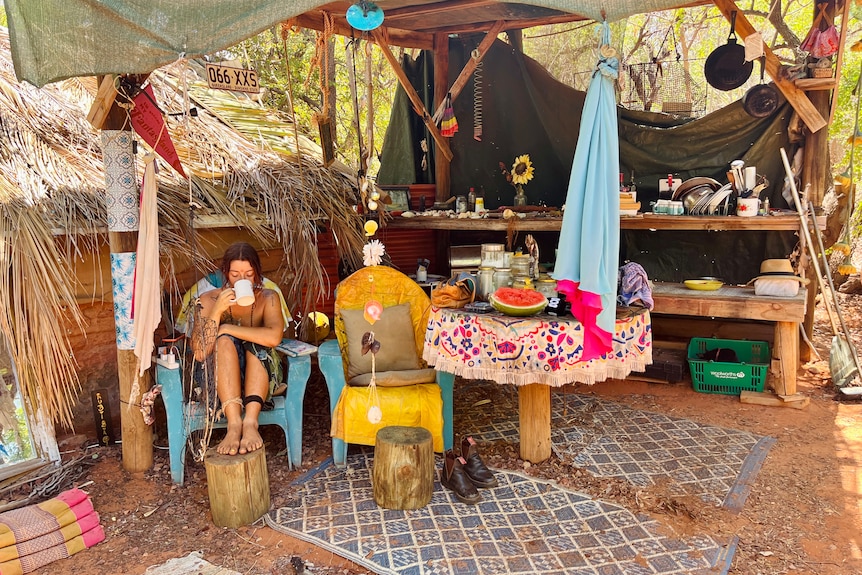 A woman sits sipping a cup of tea in an outdoor kitchen bush camp.