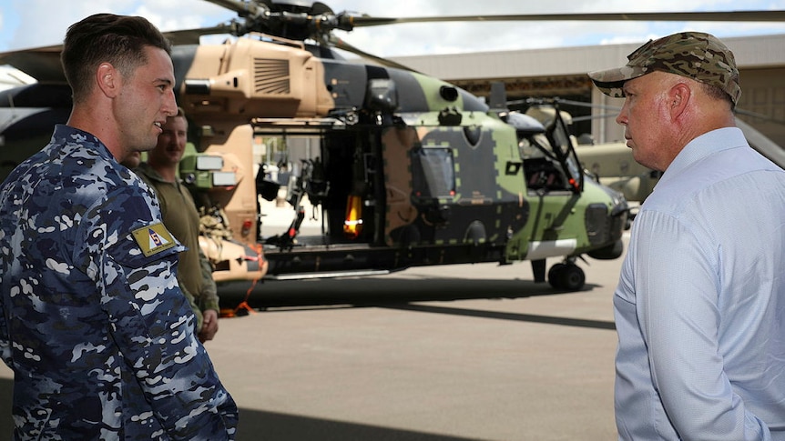 Man in navy uniform stands next man in dress shirt and cap in front of helicopter