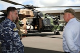 Man in navy uniform stands next man in dress shirt and cap in front of helicopter