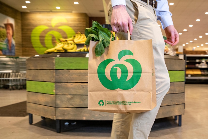 A person holds a Woolworths-branded brown paper shopping bag while walking through the fresh produce section. 