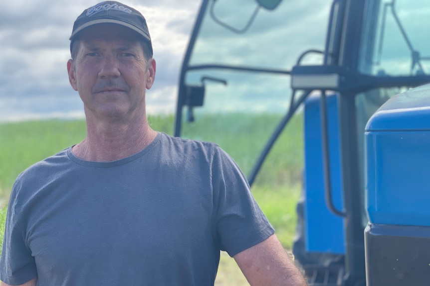 A head and shoulders portrait of a Caucasian man in a blue t-shirt and cap leaning on the front of a tractor in a cane field.