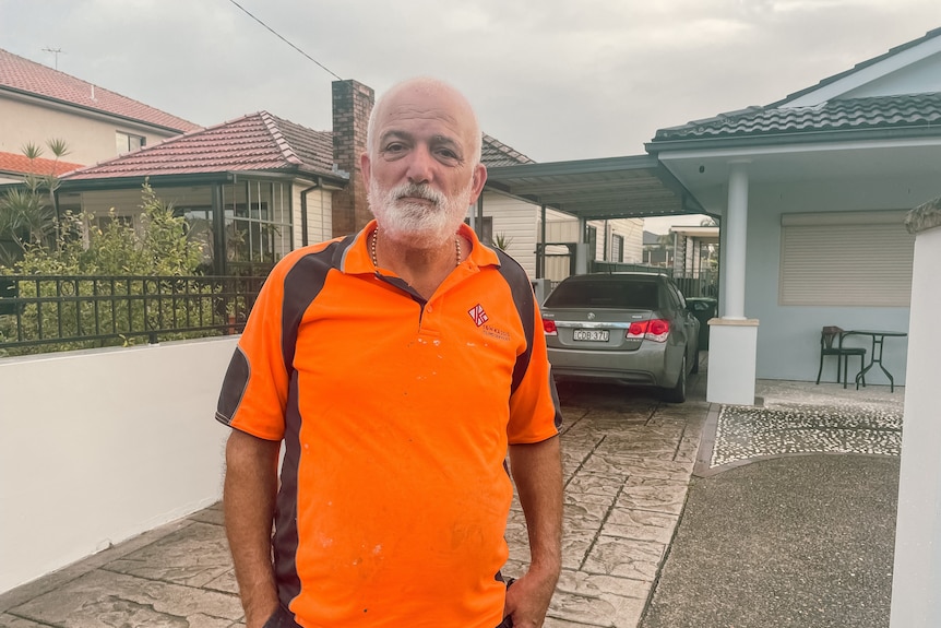A man in an orange shirt stands in front of his house