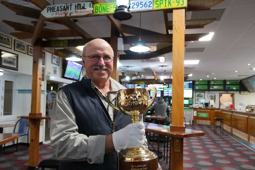 Senior man stands in pub surrounded by TV racing screens and a bar as he holds the Melbourne Cup