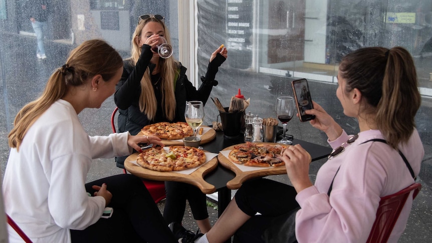 Three women sit around a table drinking wine and eating pizza at a restaurant