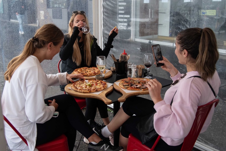 Three women sit around a table drinking wine and eating pizza at a restaurant