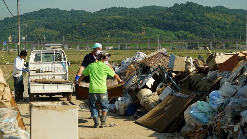 Two people carry a large item together towards a pile of rubbish.