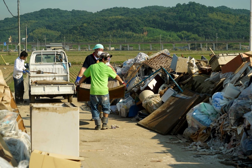 Two people carry a large item together towards a pile of rubbish.