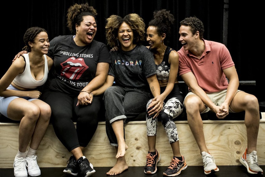 A group of actors sit on the edge of the stage and laugh. They have their arms around each or are holding hands.