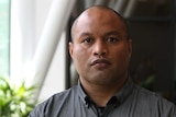 A Nauruan man with a shaved head and brown eyes looks unsmiling at the camera wearing a grey shirt.
