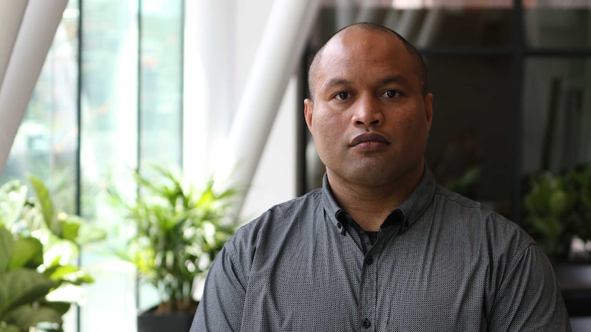 A Nauruan man with a shaved head and brown eyes looks unsmiling at the camera wearing a grey shirt.