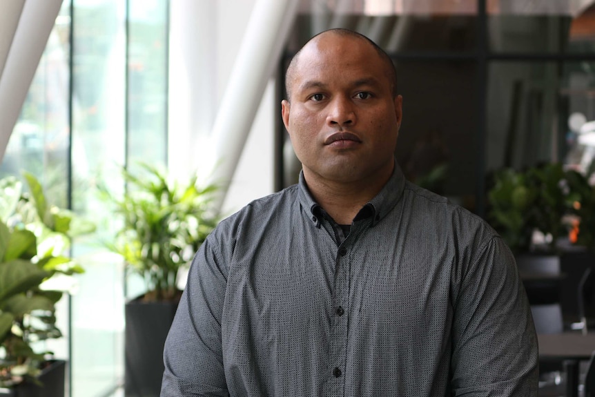 A Nauruan man with a shaved head and brown eyes looks unsmiling at the camera wearing a grey shirt.