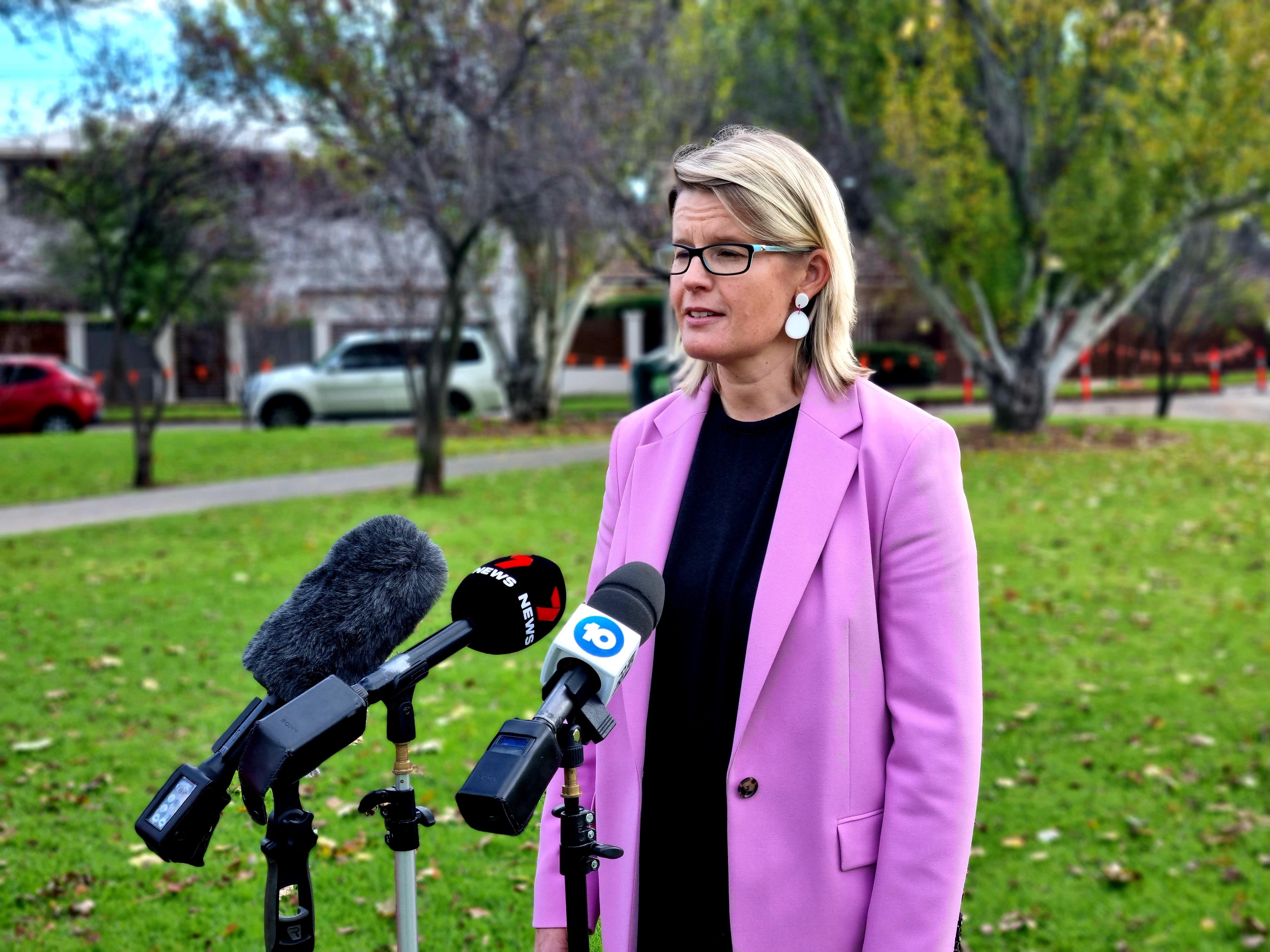 A woman with short blonde hair wearing a pink jacket stands behind microphones in a park at a media conference
