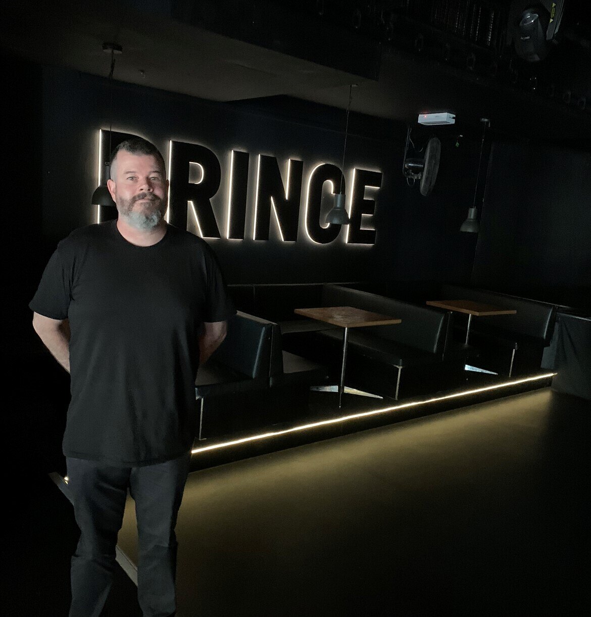 A man with a beard in a black t-shirt stands in front of booths in a venue with a lit-up sign saying 'Prince'.