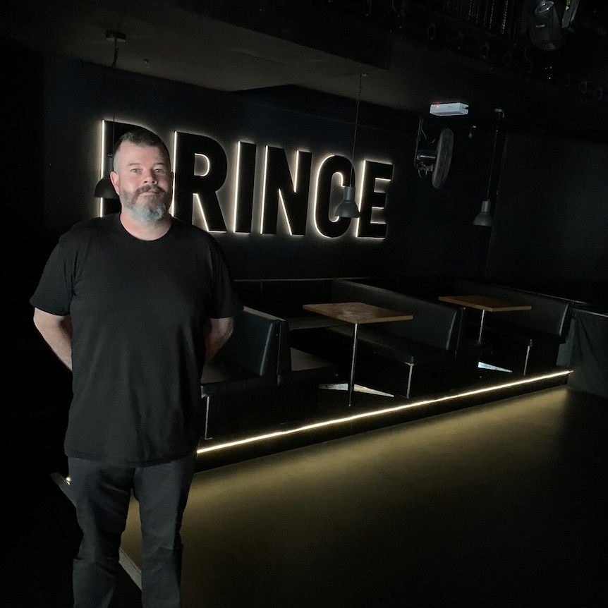 A man with a beard in a black t-shirt stands in front of booths in a venue with a lit-up sign saying 'Prince'.