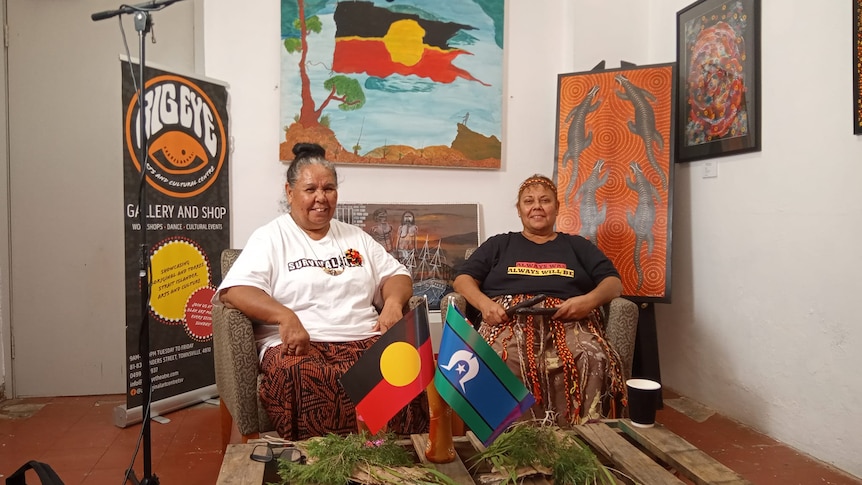 Two women sit in chairs with Aboriginal and Torres Strait Islander flags in front of them, smiling.