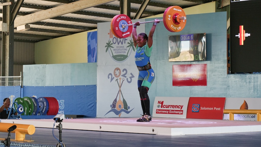 A woman lifts a large barbell over her head as she stands on a stage.