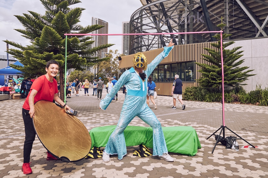 A man in a blue jumpsuit with flares and mask poses outside a football stadium, a woman in red t-shirt holds sun reflector.