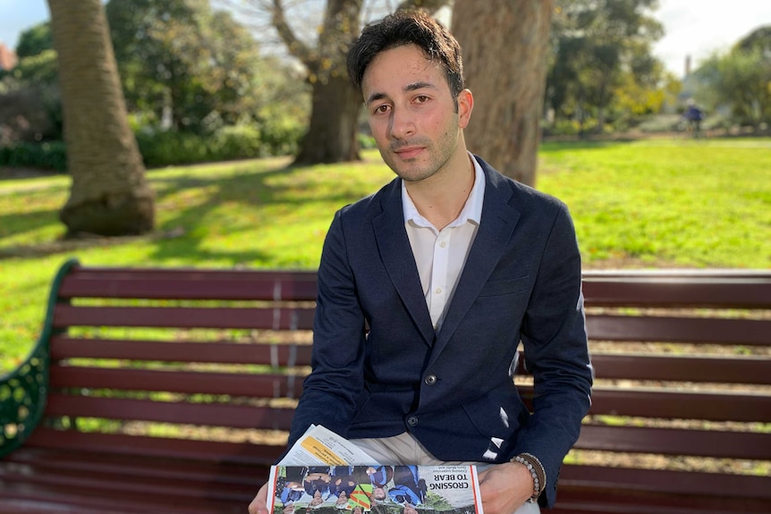 Anthony is sitting on a park bench, holding a copy of the newspaper.