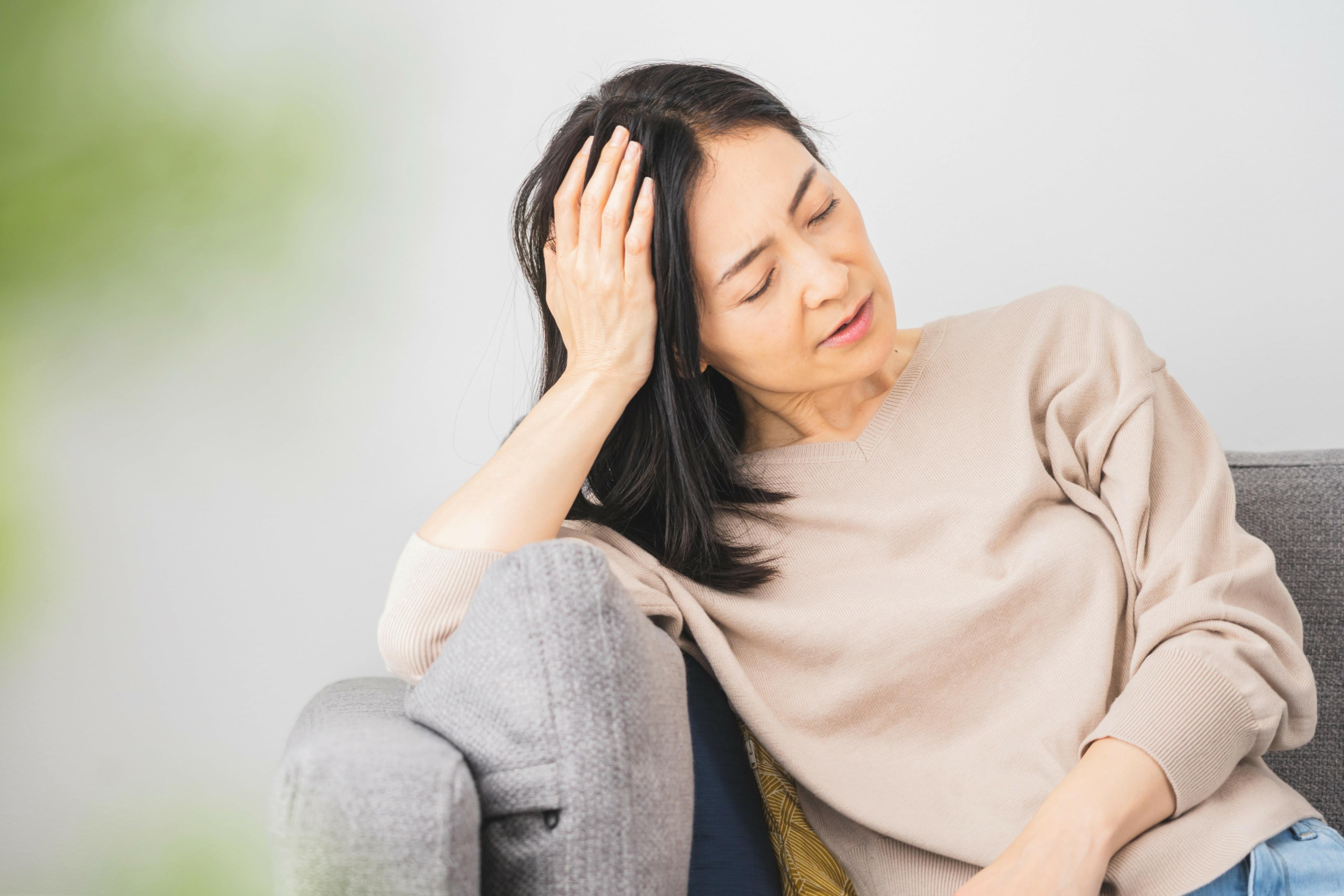 A woman with black hair closed her eyes and holds her head with her hand while sitting on a sofa.