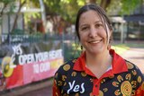 A smiling woman wearing a 'yes' shirt with for the Voice to Parliament stands in front of a 'yes' sign. Brown hair is tied back.
