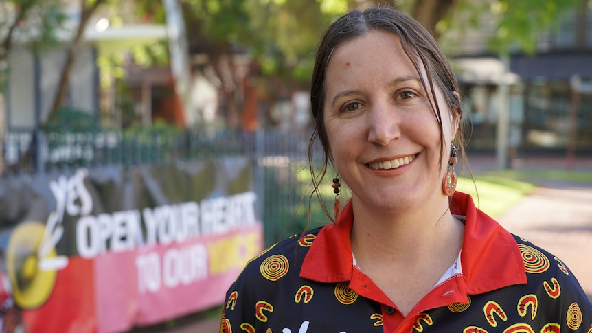 A smiling woman wearing a 'yes' shirt with for the Voice to Parliament stands in front of a 'yes' sign. Brown hair is tied back.