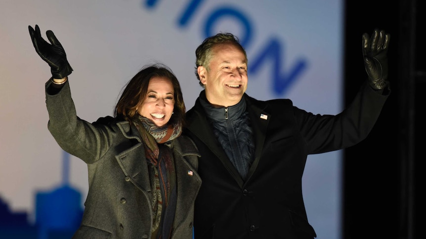 A woman and a man stand on a stage with an arm around each other and waving.