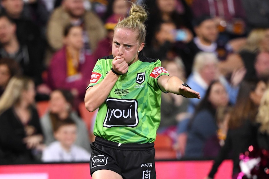 A female NRL referee blows a whistle in her right hand and points to the ground with an outstretched left arm.