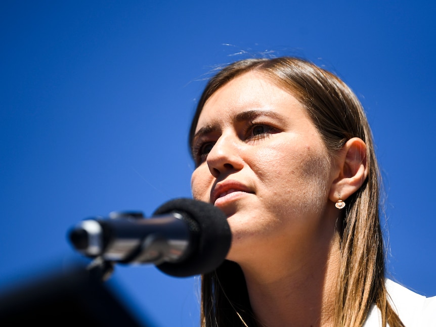 A woman with brown hair speaking into a microphone with blue sky in the background