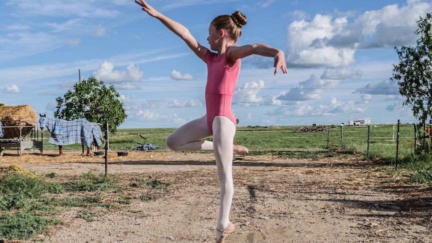 A young girl leaps in a ballet outfit in a rural setting, with horses behind her.