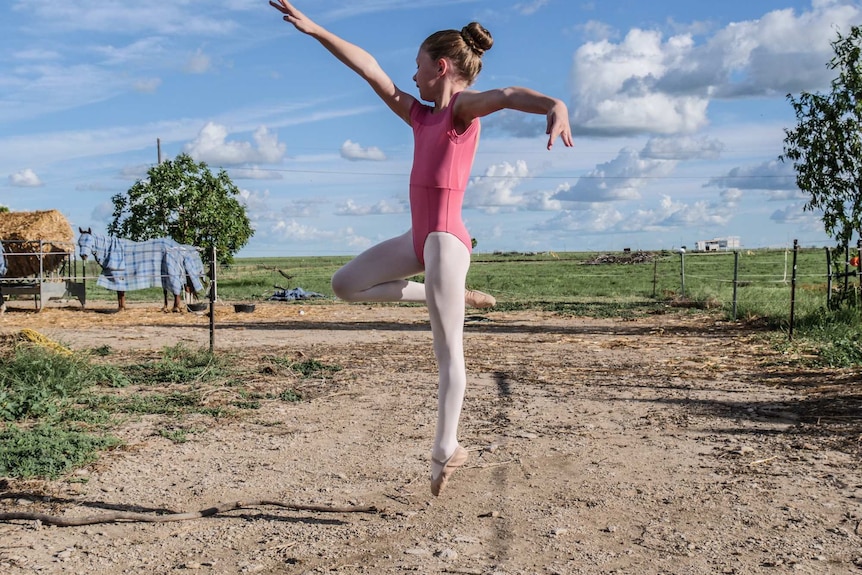 A young girl leaps in a ballet outfit in a rural setting, with horses behind her.