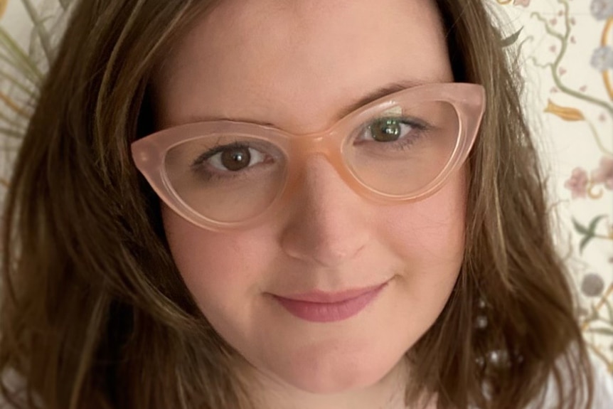 Close up of Gemma with shoulder length brown hair and 1960s style rose colored glasses smiling slightly.