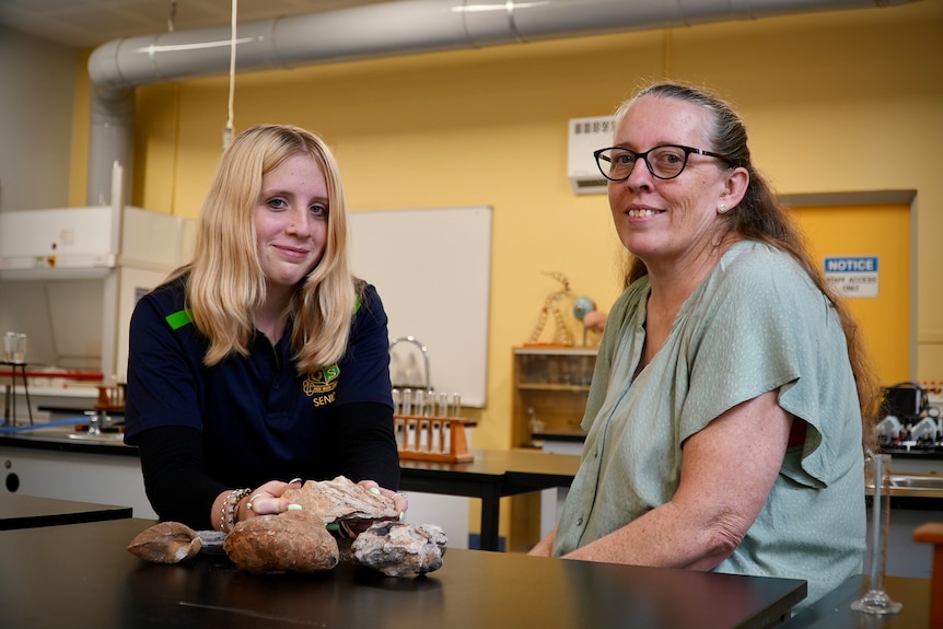 High school student with blonde hair smiles with fossil in her hand in a school science lab next to her mother.