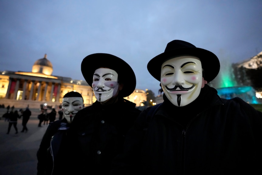 Three people wearng smiling Guy Fawkes masks pose for a photo as a crowd gathers behing them in a public square.