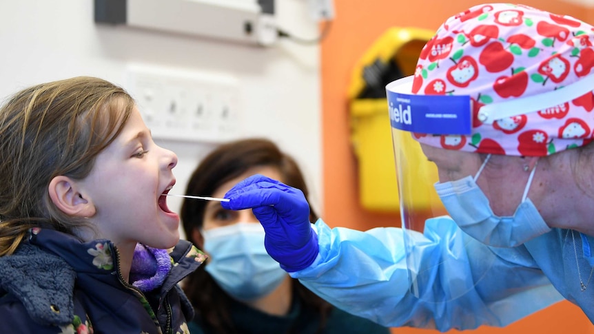 A young girl opens her mouth as a face-mask wearing doctor sticks a rod into her mouth in a clinic.