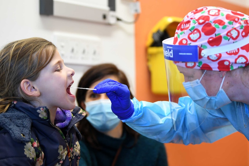 A young girl opens her mouth as a face-mask wearing doctor sticks a rod into her mouth in a clinic.
