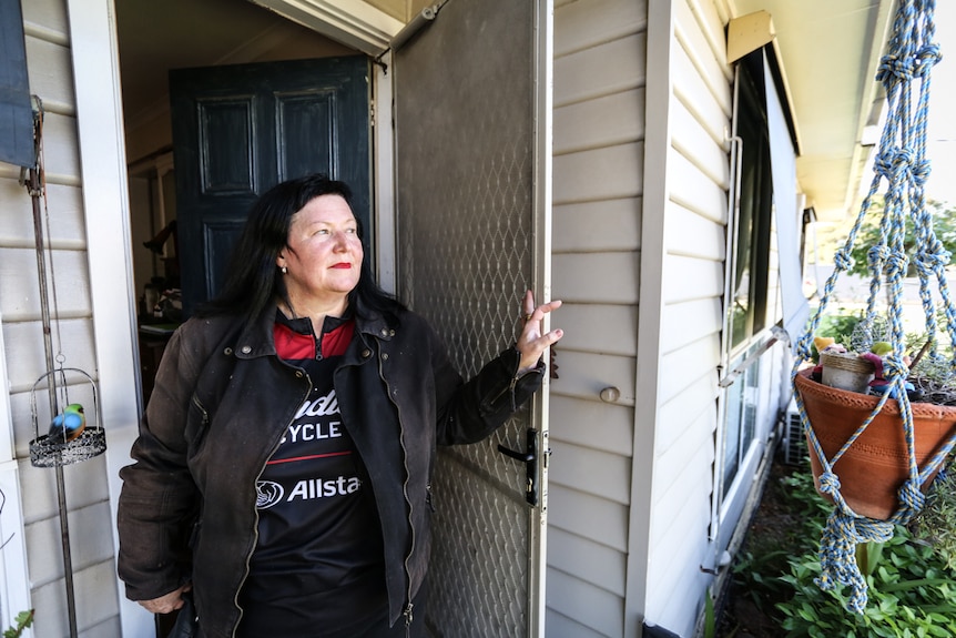 An older woman with jet-black hair and red lipstick on stands in a doorway dressed in motorcycle gear.