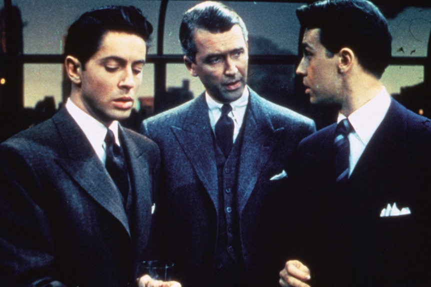 Three men in three piece grey suits stand closely together conversing in apartment with views of the city at sunset.