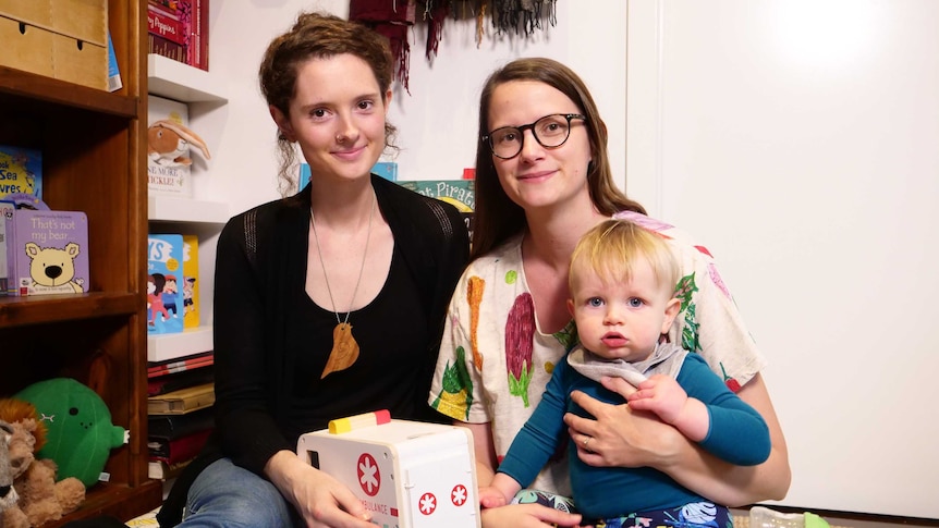 Two women smile at the camera in a play area in a home, one holds a toy ambulance, the other a male toddler.