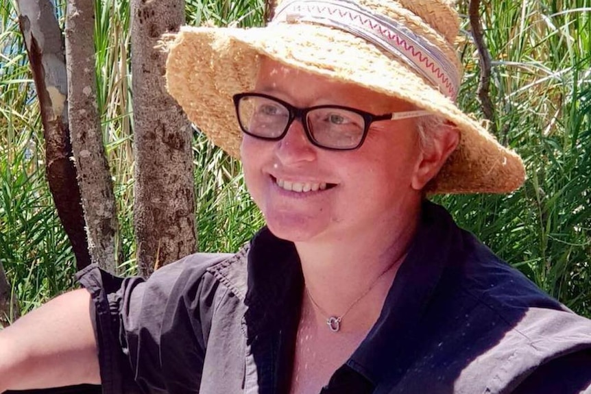 Photo of Kath O'Connor smiling widely wearing a straw hat and black-rimmed glasses. Bushland is in the background.