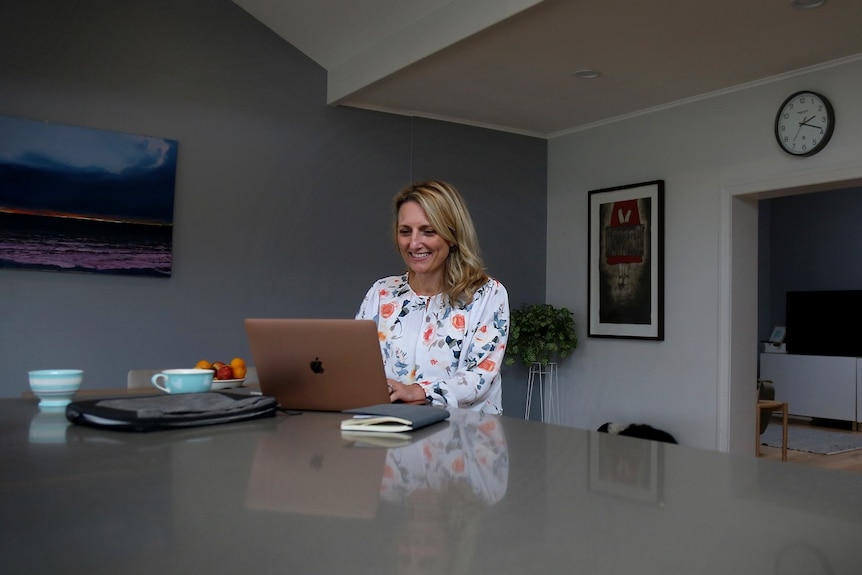 A middle-aged blonde woman sits at a kitchen table with her laptop in front of her.