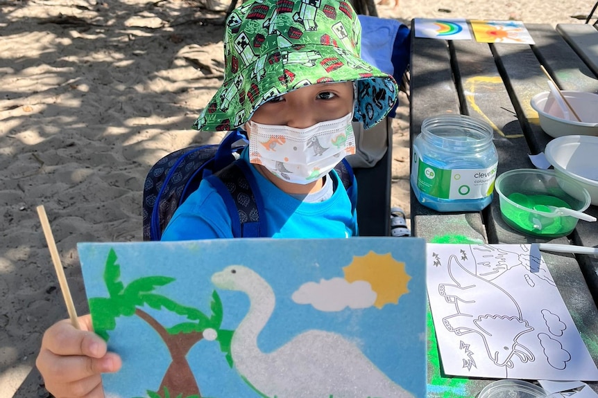 a small boy wearing a mask holds his artwork up to the camera