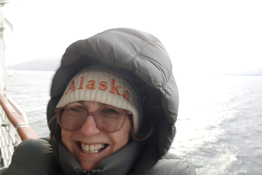 A woman wearing a beanie, puffer jacket and glasses stands on the balcony of a cruise ship