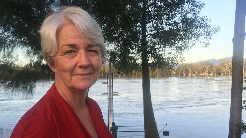 A woman with white hair and a red top stands near a river