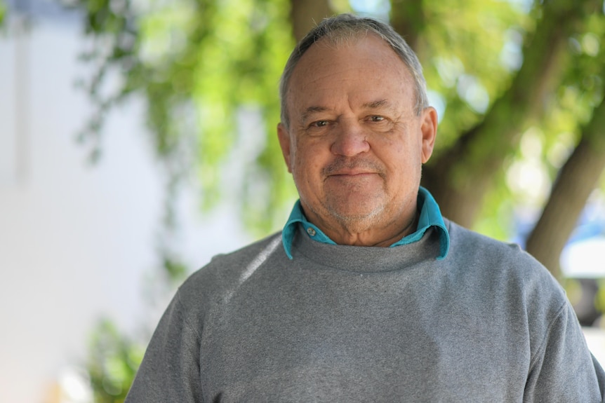 A man in a grey jumper standing in front of trees.