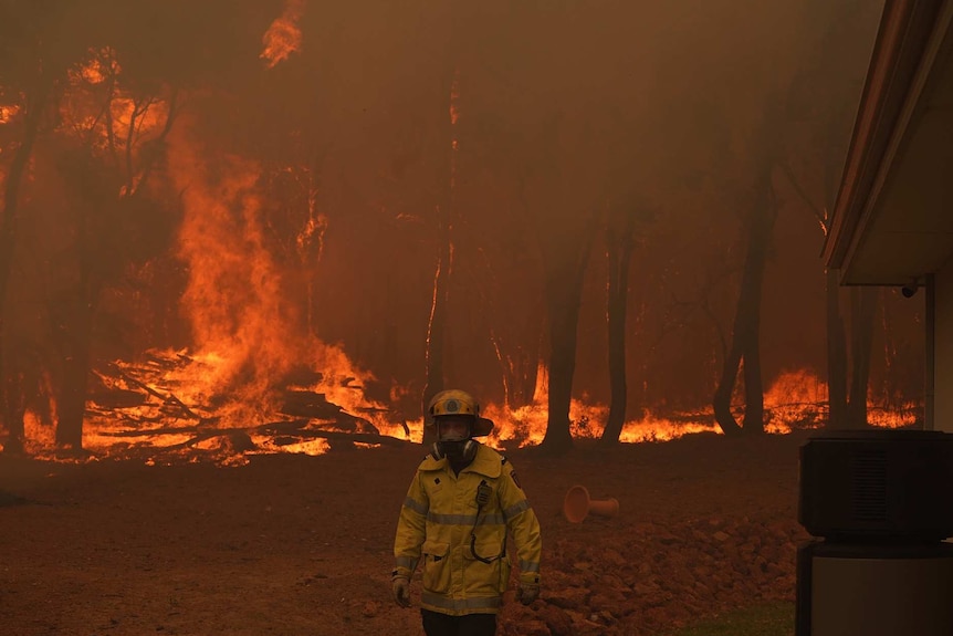 A firefighter walks away from burning bushland with a house off to his right.