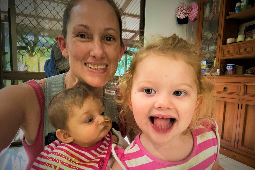 Smiling family selfie in home. Mum with baby and two-year-old. baby has a medical line to her nose.