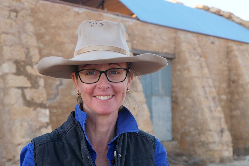 A lady wearing a weather-beaten light brown wide-brimmed hat and glasses stands in front of a woolshed made of stone.