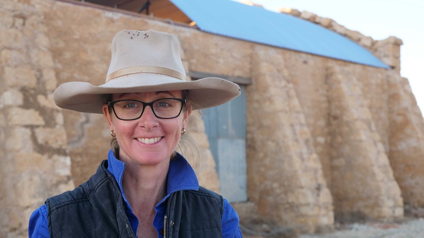 A lady wearing a weather-beaten light brown wide-brimmed hat and glasses stands in front of a woolshed made of stone.