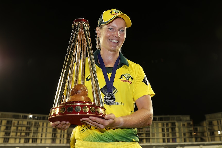 A cricketer smiles at the camera, she is holding a trophy and is wearing a medal around her neck.