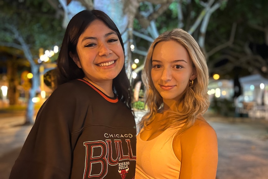 Two smiling women, one with dark hair and the other blonde, standing on a street at dusk.